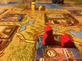 Pedine rosse su un tabellone di giochi da tavolo gestionali, con dadi e città sullo sfondo.