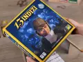 La scatola del gioco da tavolo "13 indizi" mostra un detective con lente d'ingrandimento che indaga su misteriosi personaggi in un'ambientazione notturna.