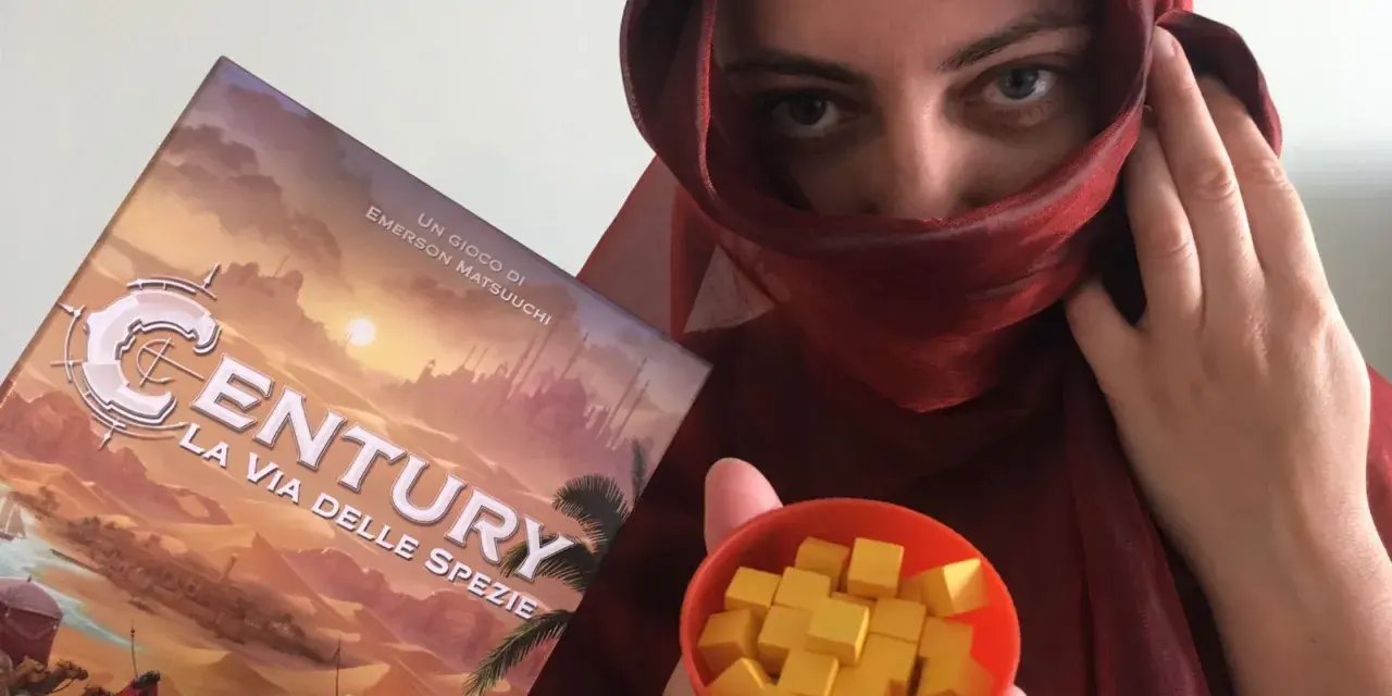 Una persona con un velo rosso tiene la scatola del gioco "Century: La via delle spezie" e una ciotola piena di cubetti gialli.