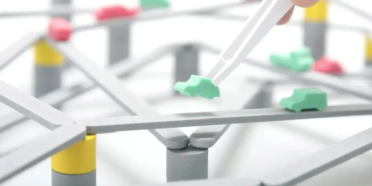 Pinzette posizionano un'auto verde su una **tokyo highway** in miniatura, con altre auto rosse e verdi sparse sulla struttura.