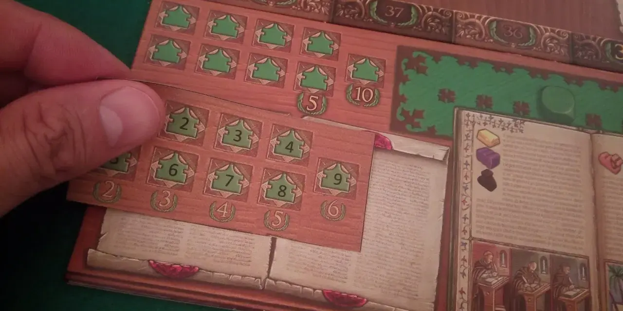 Dettaglio del gioco da tavolo "Marco Polo". Una mano posiziona una tessera con numeri verdi su una plancia di gioco in legno.