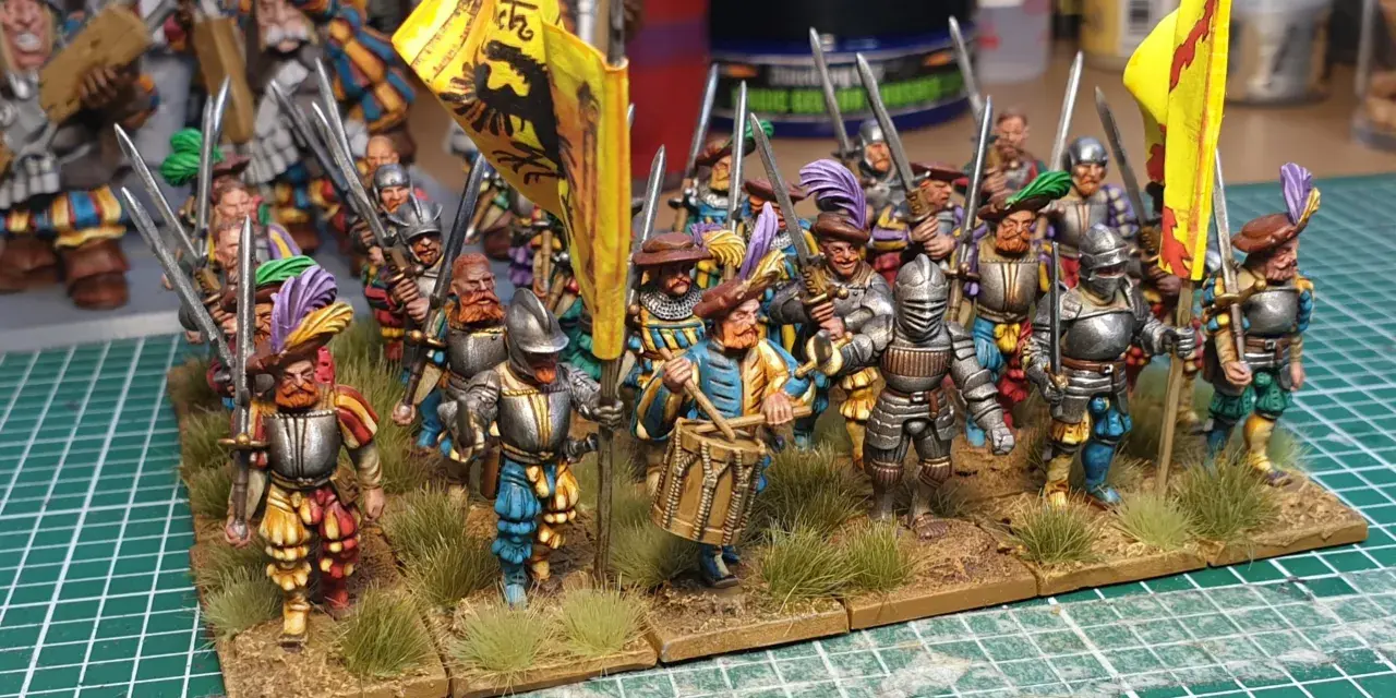 Un gruppo di soldatini in armatura, con bandiere gialle e piume colorate, pronti per la battaglia. Un tamburino guida la marcia.