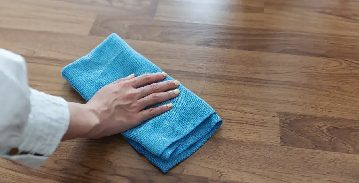 Mano con panno blu pulisce superficie in legno, mostrando come togliere la colla attak dal legno.
