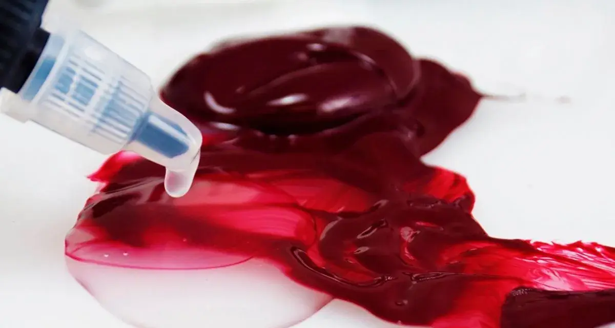Un beccuccio trasparente rilascia un liquido chiaro su un colore acrilico rosso intenso, mostrando come diluire i colori acrilici per ottenere sfumature più leggere.