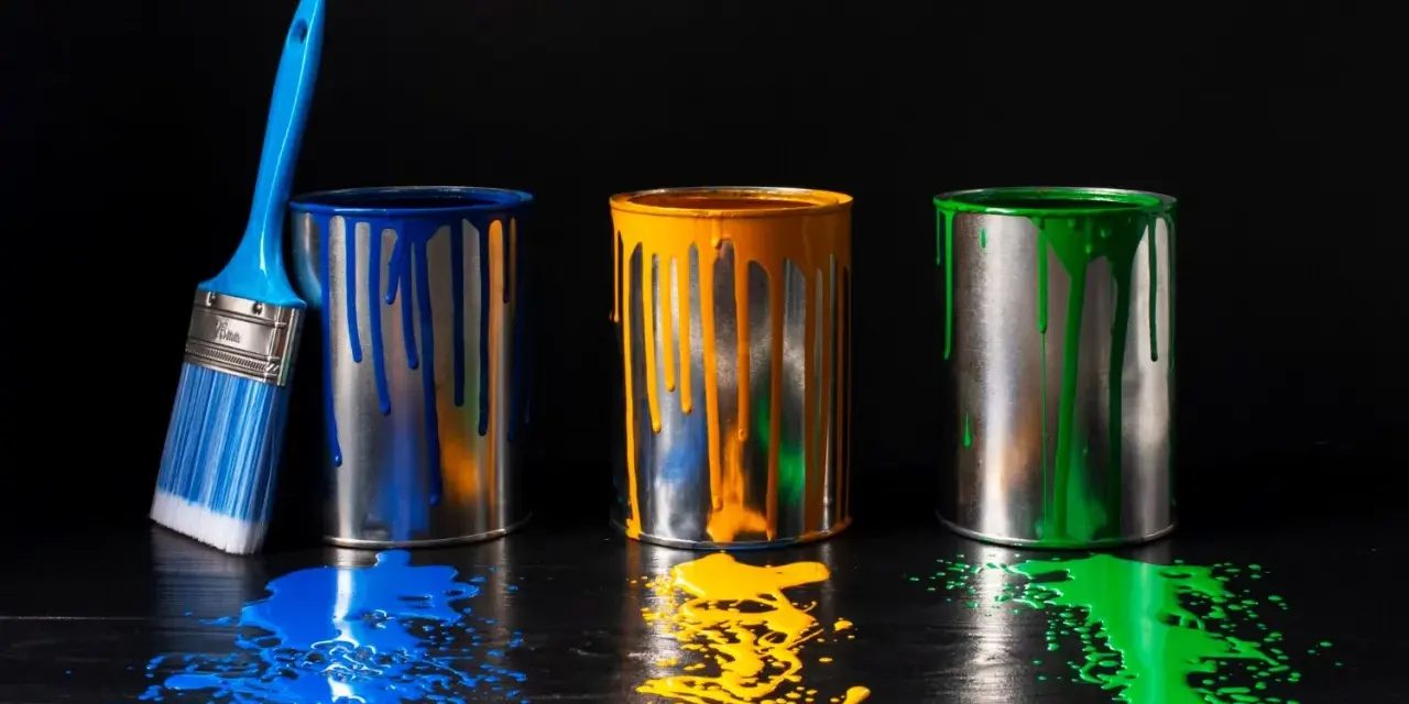 Tre barattoli di vernice blu, gialla e verde gocciolano su un tavolo di legno scuro. Un pennello blu è appoggiato al barattolo blu. Un'alternativa all'alcool isopropilico per pulire?