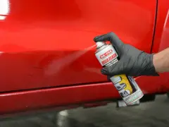 Mano con guanto nero spruzza prodotto su vernice rossa lucida, per rimuovere residui e capire come rendere opaca una vernice lucida.