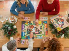 Famiglia riunita attorno a un tavolo di legno per divertenti giochi sociali, con un gioco da tavolo, carte, bevande e decorazioni natalizie.