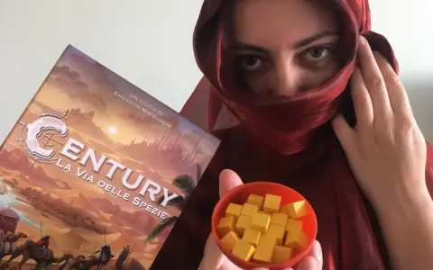 Una persona con un velo rosso tiene la scatola del gioco "Century: La via delle spezie" e una ciotola piena di cubetti gialli.