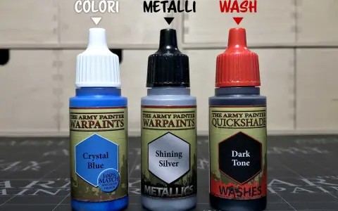 Tre boccette di colori The Army Painter: Crystal Blue (colori), Shining Silver (metalli) e Dark Tone (wash). Immagine con colore desaturato.
