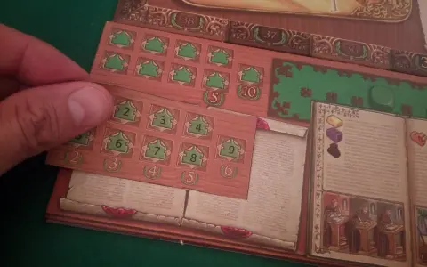 Dettaglio del gioco da tavolo "Marco Polo". Una mano posiziona una tessera con numeri verdi su una plancia di gioco in legno.