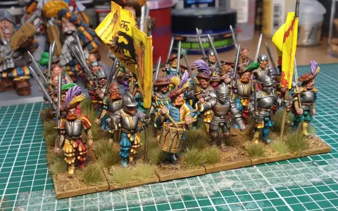 Un gruppo di soldatini in armatura, con bandiere gialle e piume colorate, pronti per la battaglia. Un tamburino guida la marcia.
