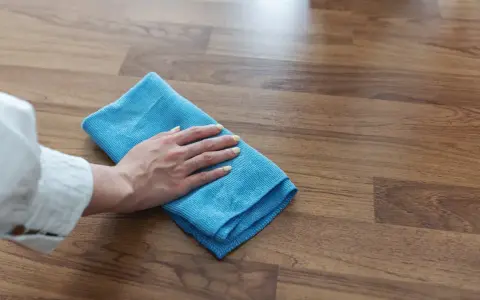Mano con panno blu pulisce superficie in legno, mostrando come togliere la colla attak dal legno.