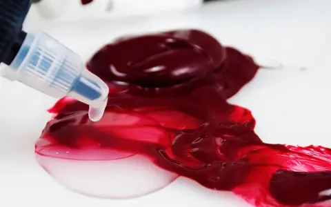 Un beccuccio trasparente rilascia un liquido chiaro su un colore acrilico rosso intenso, mostrando come diluire i colori acrilici per ottenere sfumature più leggere.