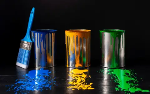 Tre barattoli di vernice blu, gialla e verde gocciolano su un tavolo di legno scuro. Un pennello blu è appoggiato al barattolo blu. Un'alternativa all'alcool isopropilico per pulire?