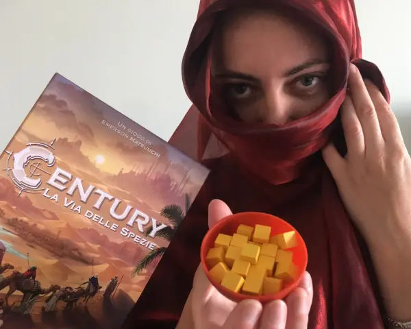 Una persona con un velo rosso tiene la scatola del gioco "Century: La via delle spezie" e una ciotola piena di cubetti gialli.