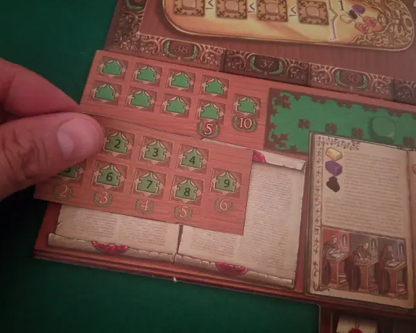 Dettaglio del gioco da tavolo "Marco Polo". Una mano posiziona una tessera con numeri verdi su una plancia di gioco in legno.
