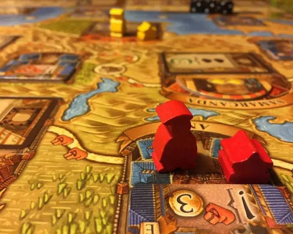 Pedine rosse su un tabellone di giochi da tavolo gestionali, con dadi e città sullo sfondo.