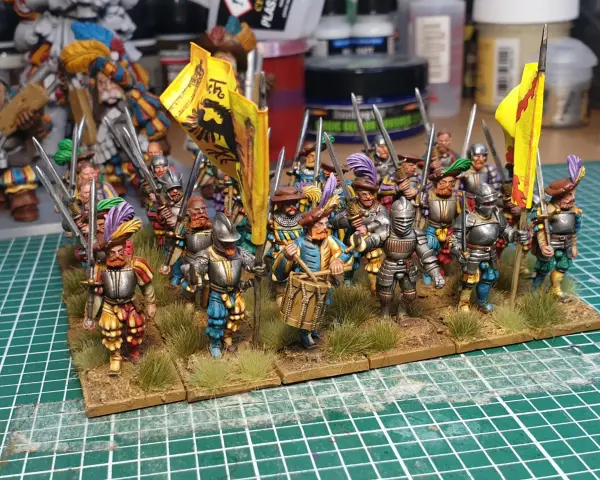Un gruppo di soldatini in armatura, con bandiere gialle e piume colorate, pronti per la battaglia. Un tamburino guida la marcia.