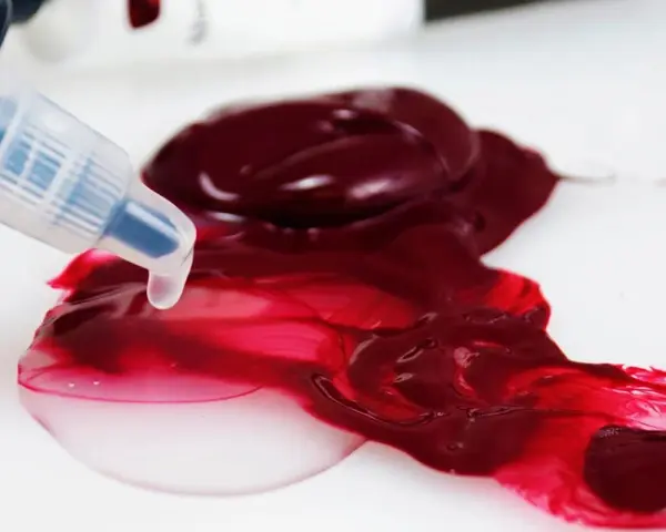 Un beccuccio trasparente rilascia un liquido chiaro su un colore acrilico rosso intenso, mostrando come diluire i colori acrilici per ottenere sfumature più leggere.