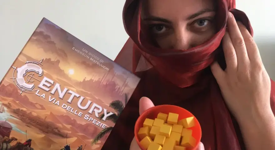 Una persona con un velo rosso tiene la scatola del gioco "Century: La via delle spezie" e una ciotola piena di cubetti gialli.