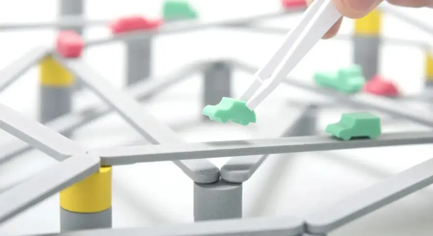 Pinzette posizionano un'auto verde su una **tokyo highway** in miniatura, con altre auto rosse e verdi sparse sulla struttura.