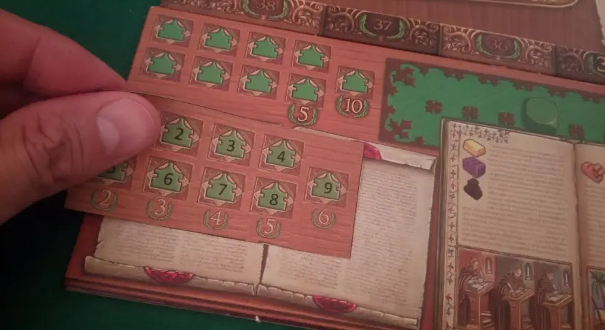 Dettaglio del gioco da tavolo "Marco Polo". Una mano posiziona una tessera con numeri verdi su una plancia di gioco in legno.