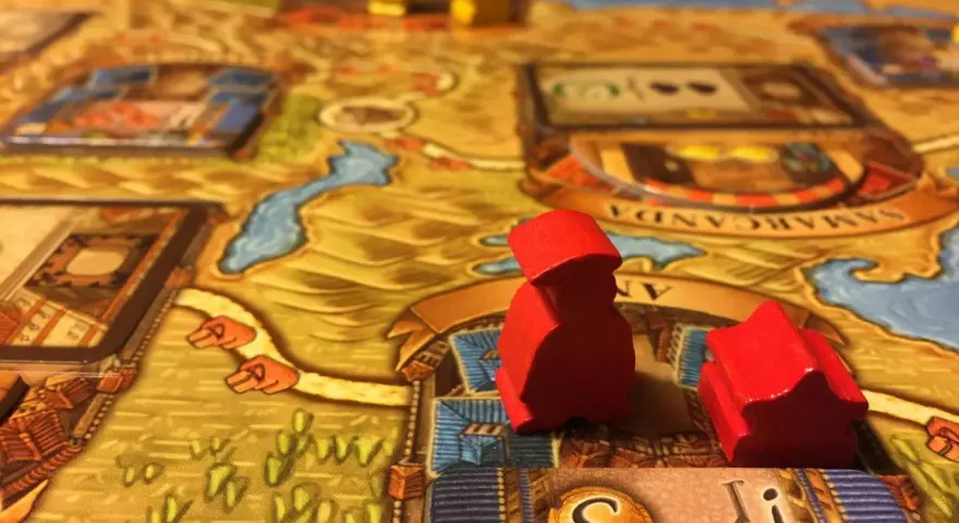 Pedine rosse su un tabellone di giochi da tavolo gestionali, con dadi e città sullo sfondo.