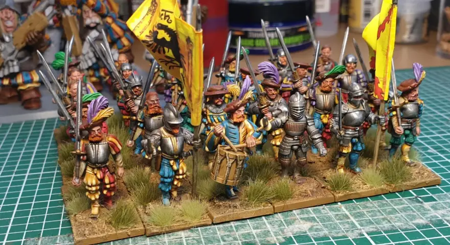 Un gruppo di soldatini in armatura, con bandiere gialle e piume colorate, pronti per la battaglia. Un tamburino guida la marcia.