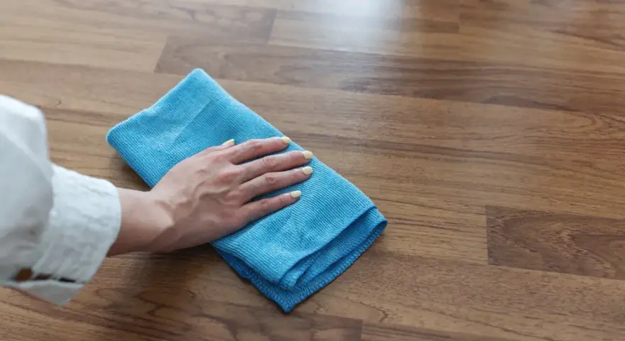 Mano con panno blu pulisce superficie in legno, mostrando come togliere la colla attak dal legno.