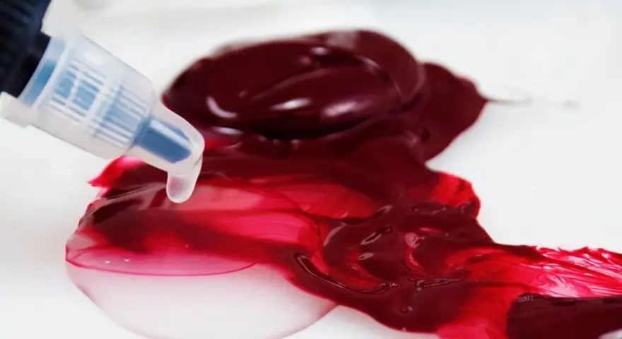 Un beccuccio trasparente rilascia un liquido chiaro su un colore acrilico rosso intenso, mostrando come diluire i colori acrilici per ottenere sfumature più leggere.