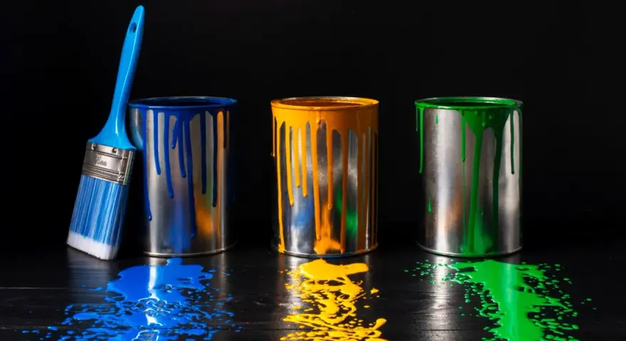 Tre barattoli di vernice blu, gialla e verde gocciolano su un tavolo di legno scuro. Un pennello blu è appoggiato al barattolo blu. Un'alternativa all'alcool isopropilico per pulire?