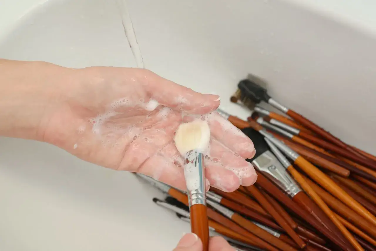 Mano che lava un pennello da trucco con sapone sotto l'acqua corrente. Molti altri pennelli sono appoggiati nel lavandino.