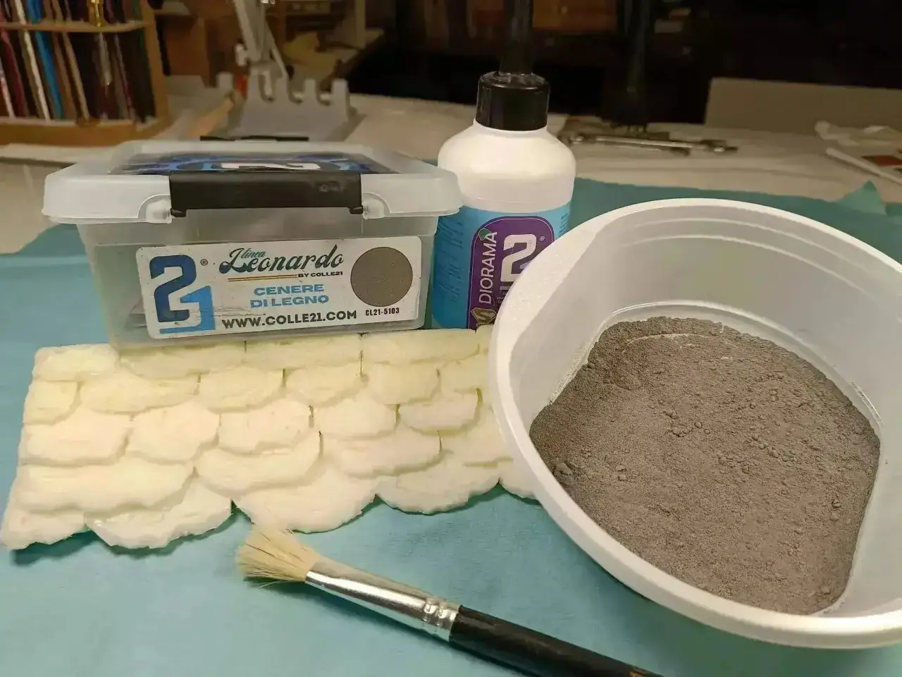 Materiali per diorami: sabbia e colla vinilica pronti per creare texture realistiche. Pennello e base in schiuma completano la scena.