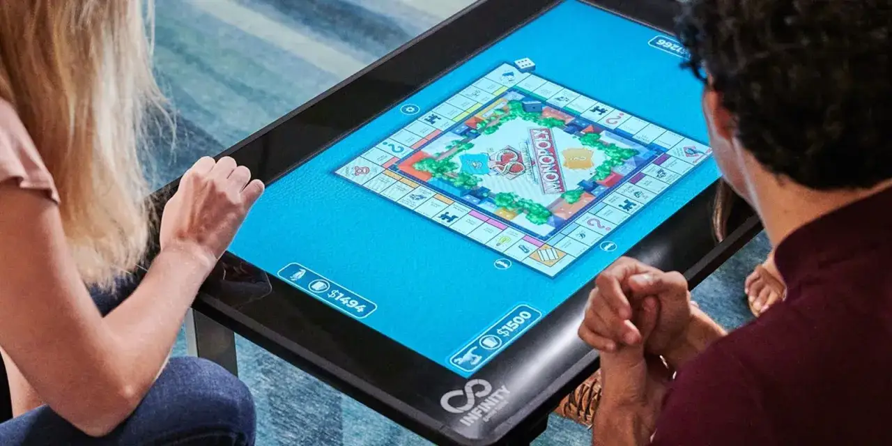Coppia gioca a Monopoly su un tavolo interattivo. 13 indizi per vincere la partita.