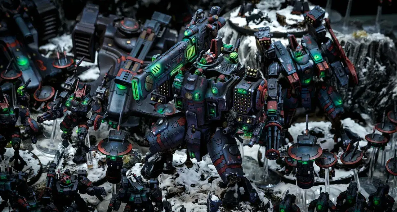 Un imponente mech tau 40k domina la scena, circondato da unit&agrave; pi&ugrave; piccole e veicoli da guerra su un terreno innevato.