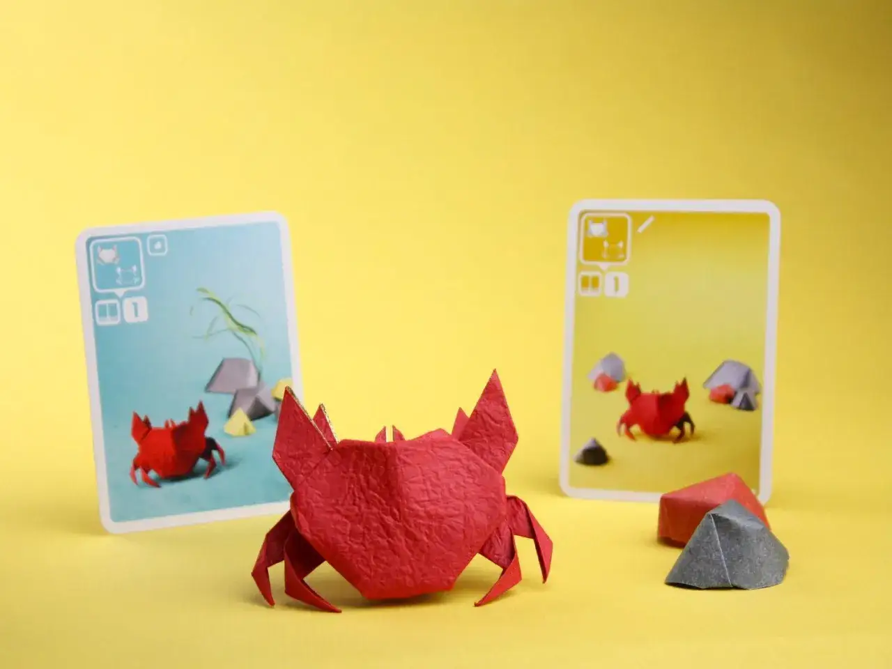 Granchi rossi di carta su sfondo giallo, con carte gioco che raffigurano granchi e rocce. Un tocco di sale marino e carta per un gioco da tavolo.