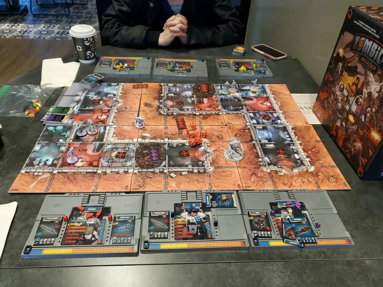 Zombicide Invader: una partita in corso con miniature, dadi rossi e carte. La scatola del gioco &egrave; visibile.