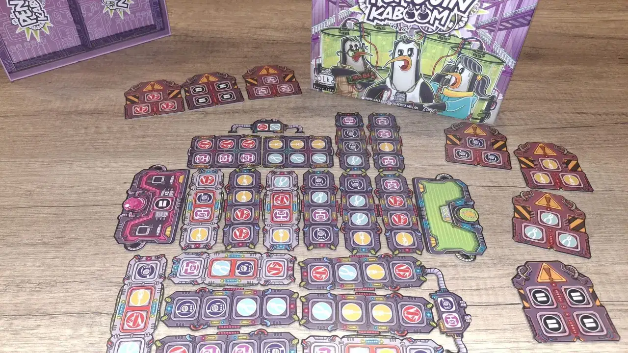 Componenti del gioco di carte Kaboom sparsi su un tavolo di legno. I pezzi colorati raffigurano pinguini e vari strumenti.