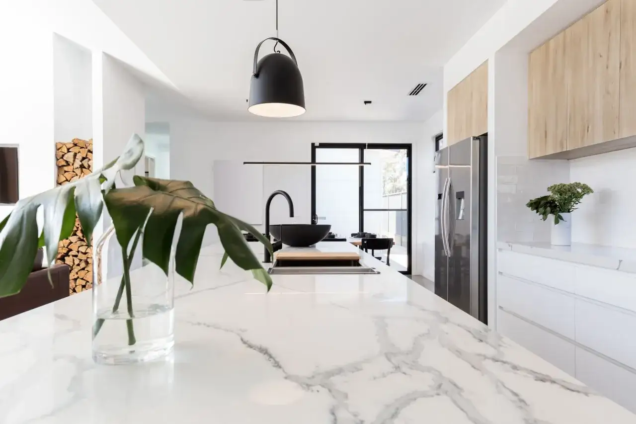Isola cucina moderna con piano in marmo e lavello nero. Sfumature di bianco dominano l'ambiente, con dettagli in legno e piante verdi.