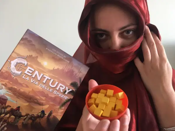 Una persona con un velo rosso tiene la scatola del gioco "Century: La via delle spezie" e una ciotola piena di cubetti gialli.