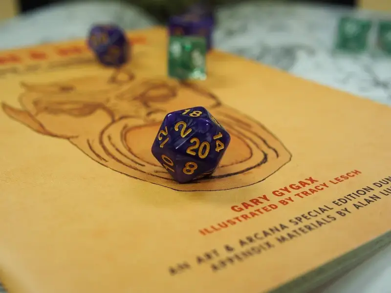 Dadi viola e verdi pronti per un'avventura di gioco di ruolo. Il d20 è in primo piano su un libro con un disegno stilizzato.